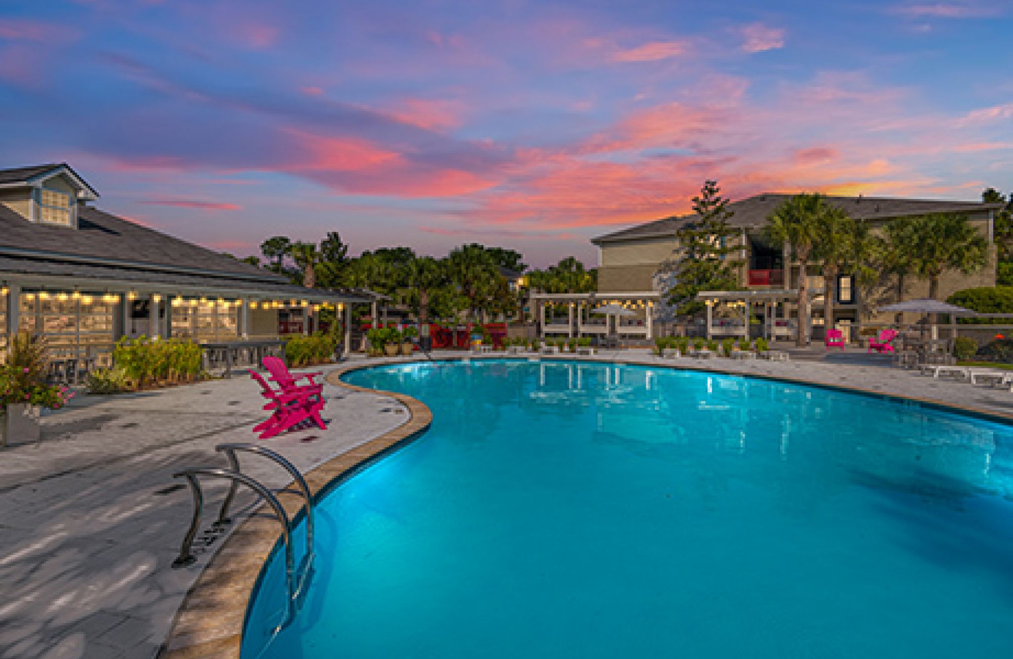 A serene outdoor pool at Atlantic Palms, surrounded by lounge chairs and buildings at sunset, with a colorful sky reflecting in the water.