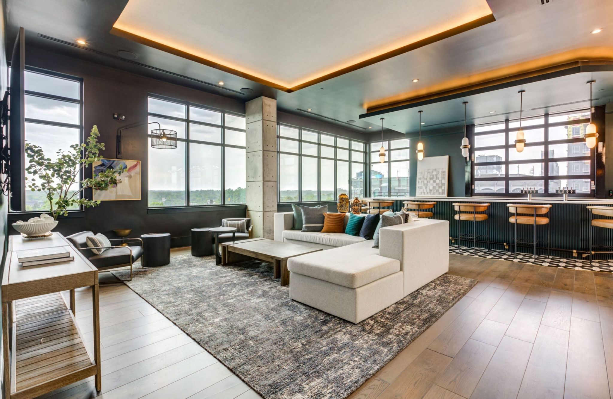 Modern open-concept living space with gray sectional sofa, bar area, large windows, and decorative lighting.