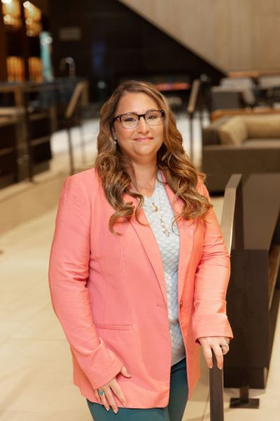 Person in glasses wearing a pink blazer stands in a modern lobby.