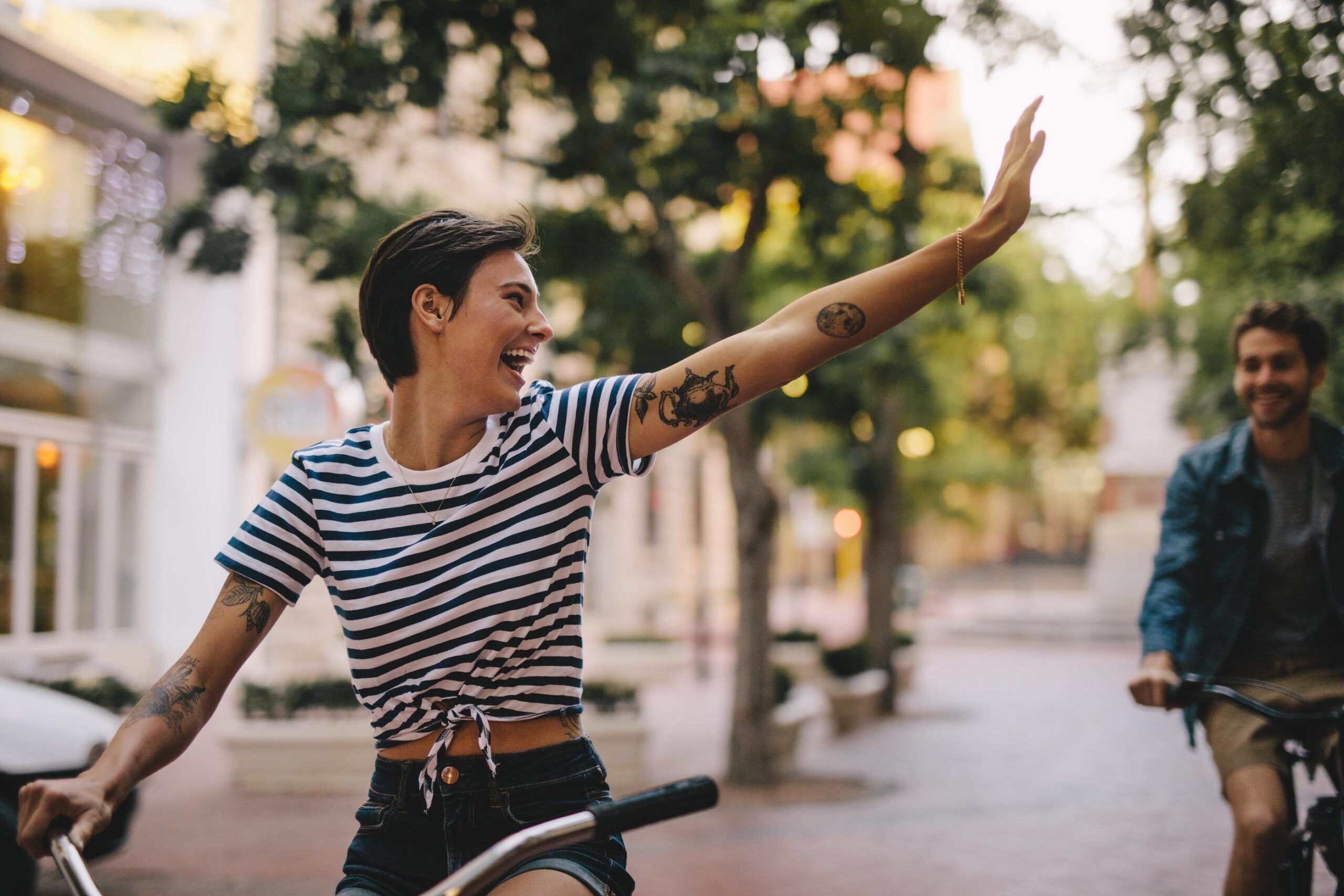 A person in a striped shirt and vest rides a bike, smiling and waving as they cruise down a tree-lined street in a charming residential area.