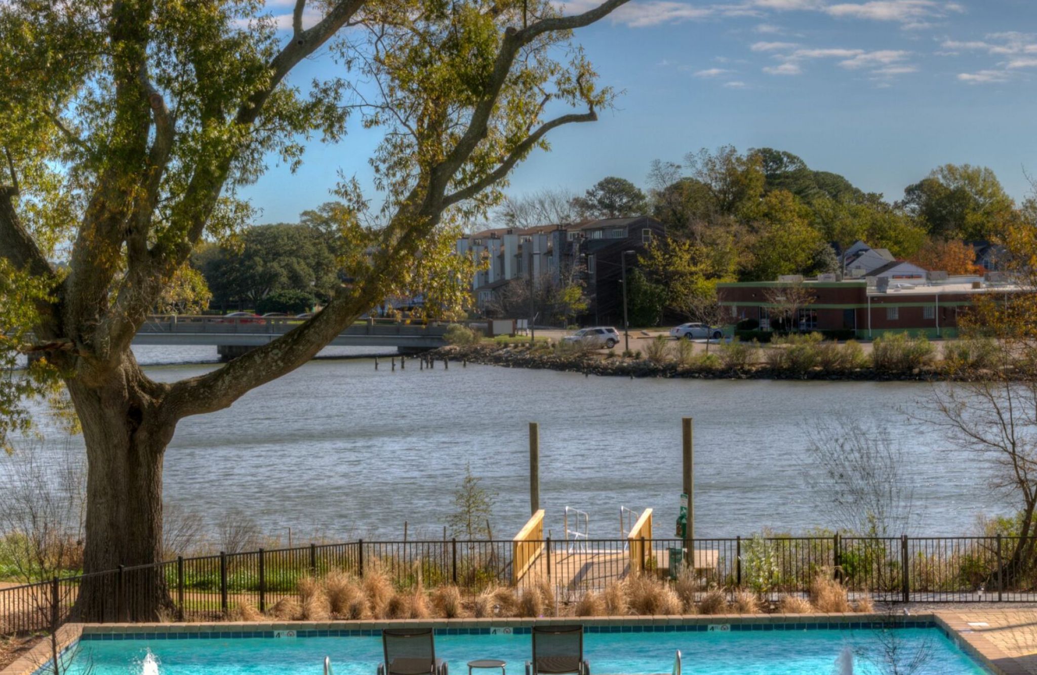 View of a pool at CovePointe with lounge chairs, a tree, and a river under the partly cloudy sky.