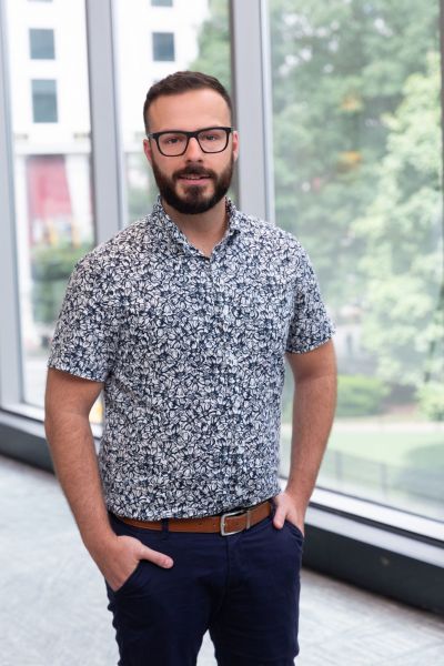 Man with glasses in a patterned shirt stands by large windows with a view of trees and buildings.