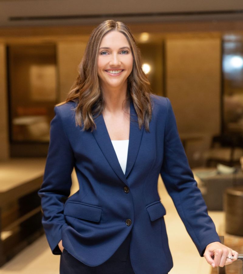 Smiling woman in a blue suit standing indoors with one hand in her pocket.