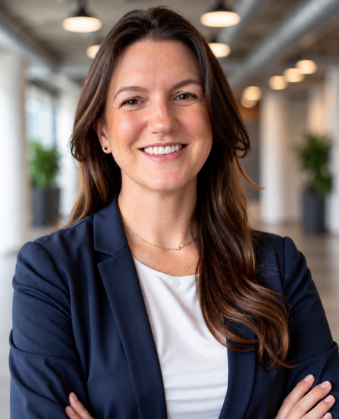 Victoria Pakala, smiling in a navy blazer, stands with arms crossed in a bright, modern office hallway.