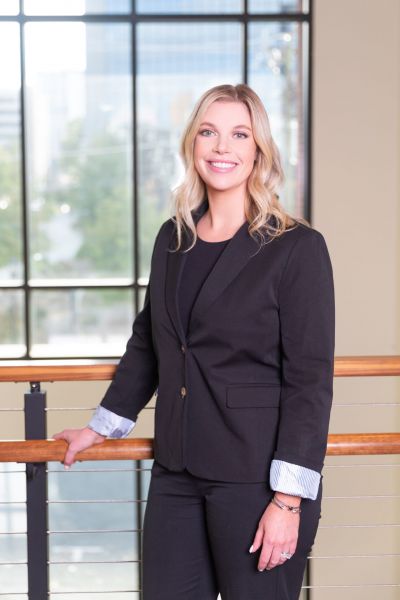 Person in a black suit standing by a railing, smiling, with large windows and a cityscape in the background.