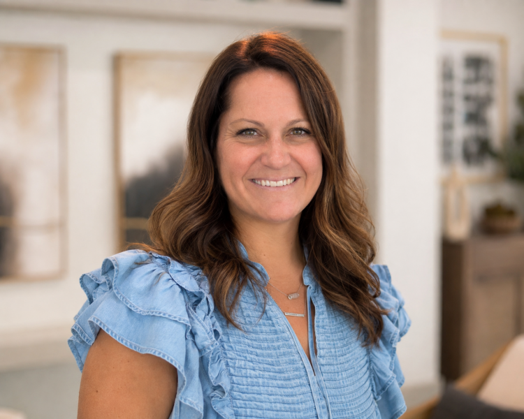Miranda Hooghkirk, with wavy brown hair, wears a blue ruffled blouse and smiles indoors with abstract art in the background.