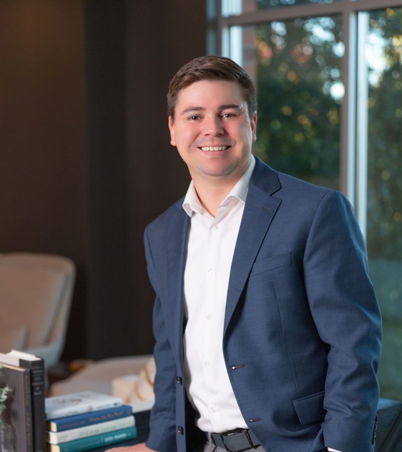 Person in a blue blazer smiles while leaning on a table with books in a well-lit room.