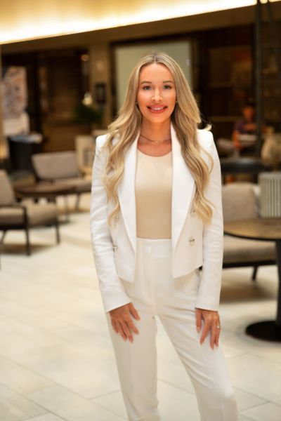 A woman in a white suit stands in a modern, well-lit indoor setting with tables and chairs in the background.