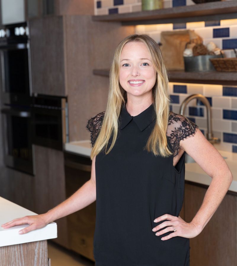 Person smiling in a modern kitchen, wearing a black top with lace sleeves, standing by a countertop.