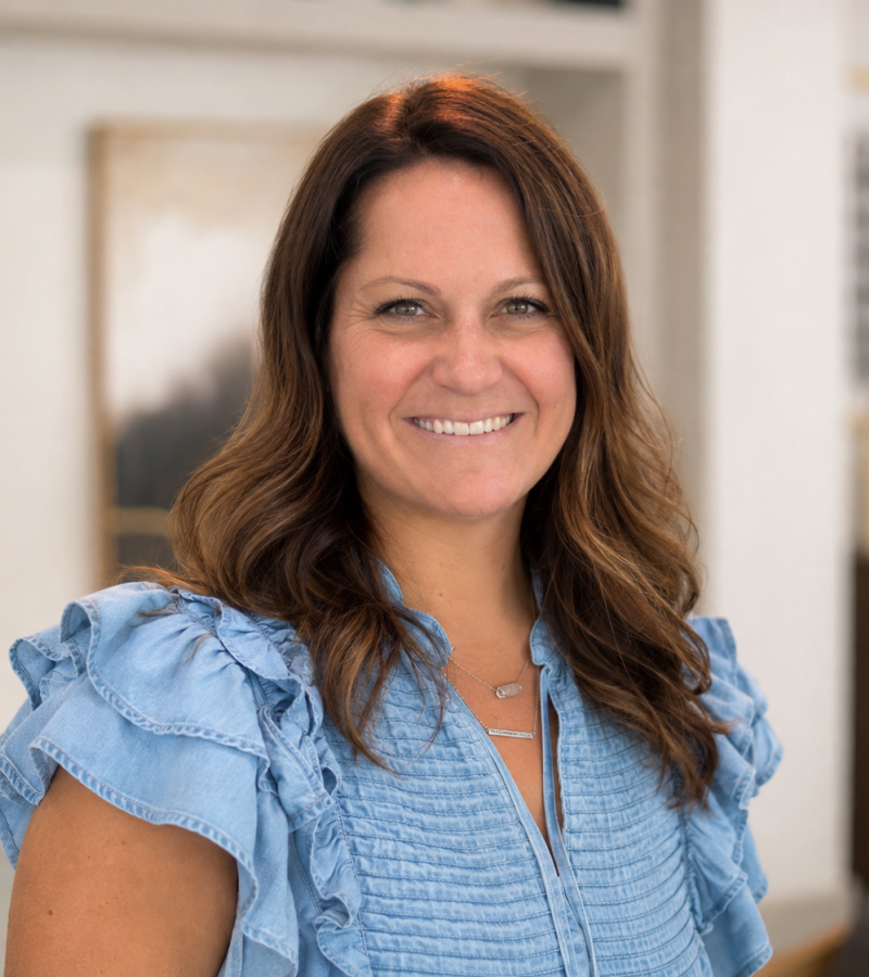 Miranda Hooghkirk, with wavy brown hair, wears a blue ruffled blouse and smiles indoors with abstract art in the background.