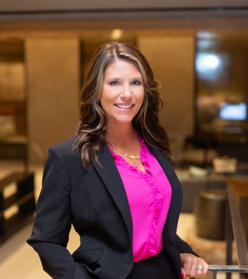 A woman in a black suit and pink blouse stands smiling in a modern office setting.