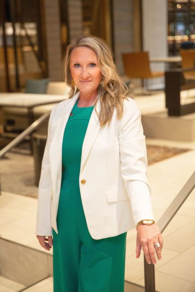 Woman in white blazer and green outfit standing indoors near stairs, looking at the camera.