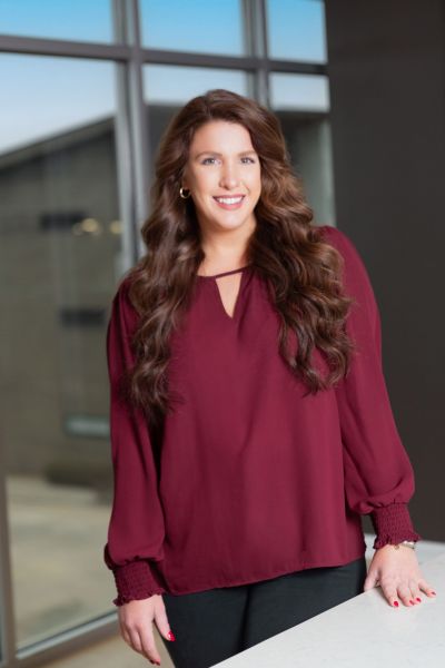 Smiling woman in a maroon blouse stands by a table in a modern office with large windows.