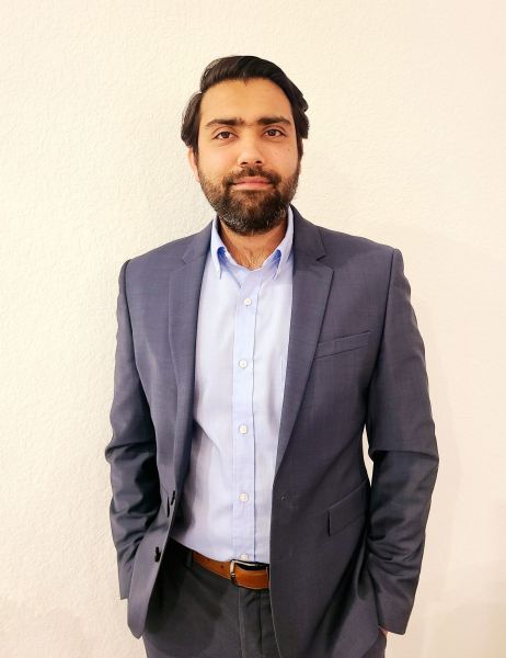 A man in a blue suit and light blue shirt stands in front of a white wall, looking at the camera.