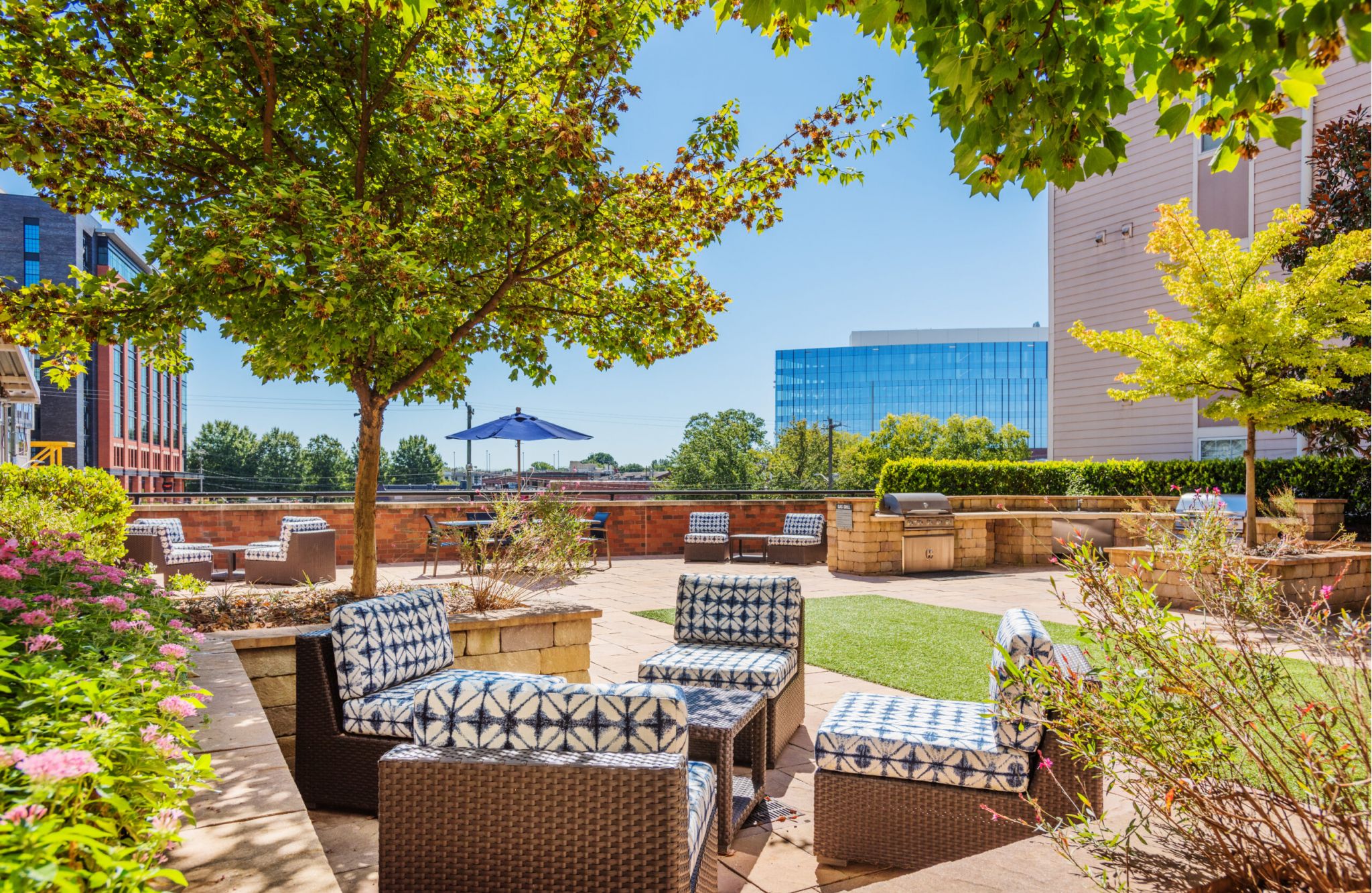 Outdoor patio at District Flats with patterned chairs, tables, a blue umbrella, and greenery. Buildings stand tall under a clear sky.