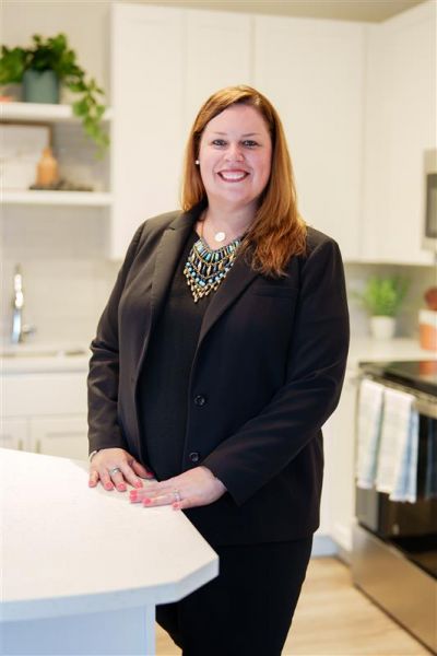 Smiling woman in a black suit, Ashley Dugger, stands in a modern kitchen with white cabinets and plants.