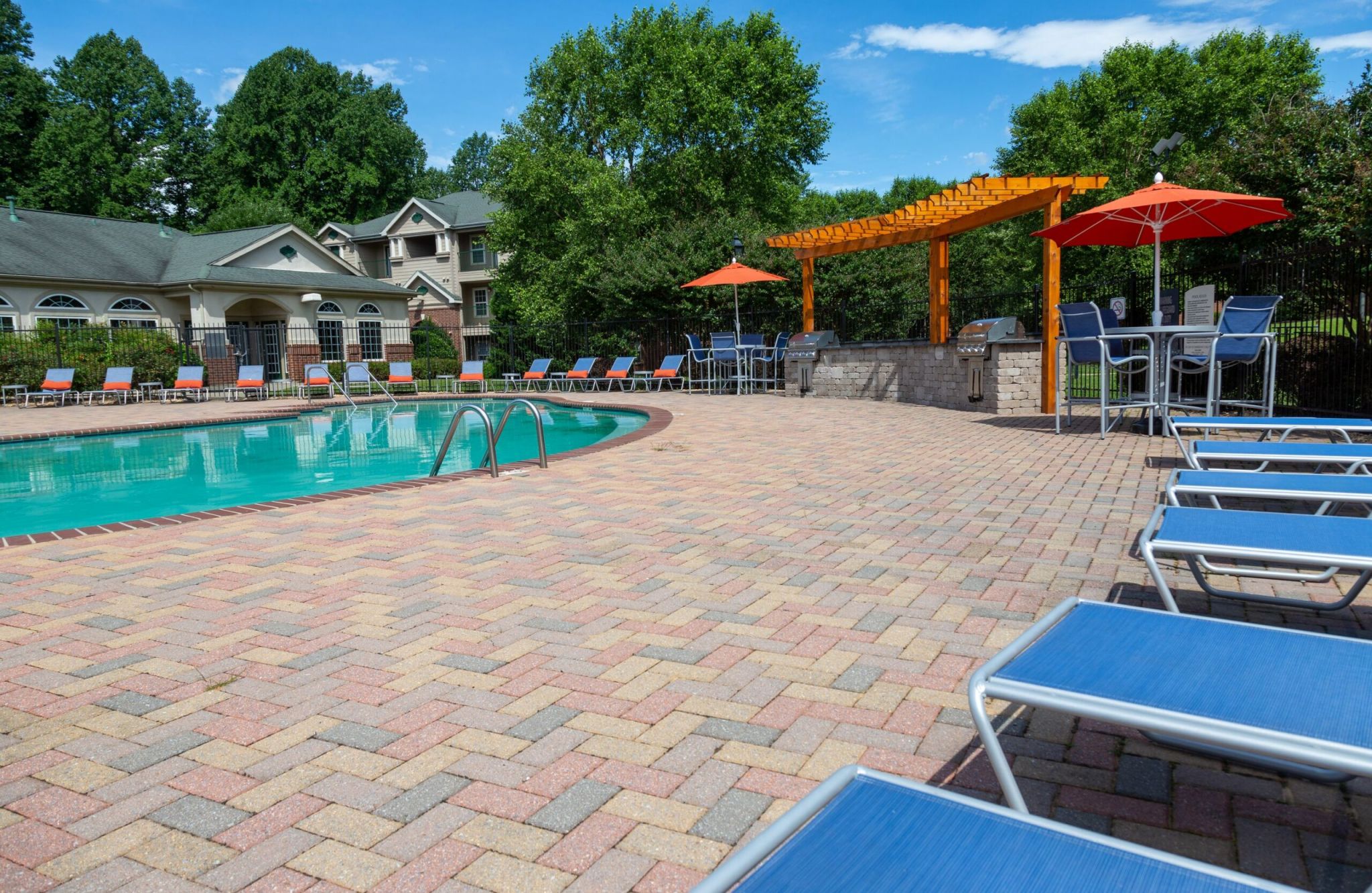 A sunny 5th-story oasis with an outdoor pool, lounge chairs, a pergola, and umbrellas, all surrounded by lush greenery and nearby buildings.