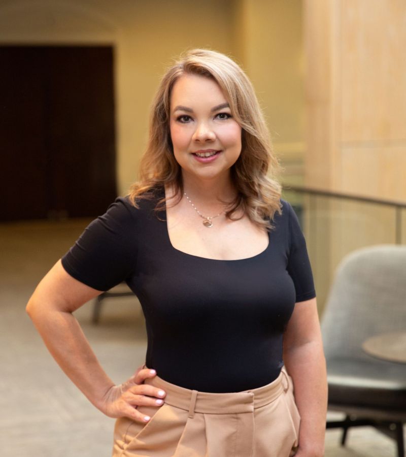 Smiling woman in a black top and beige pants stands in a modern indoor setting with a hand on her hip.