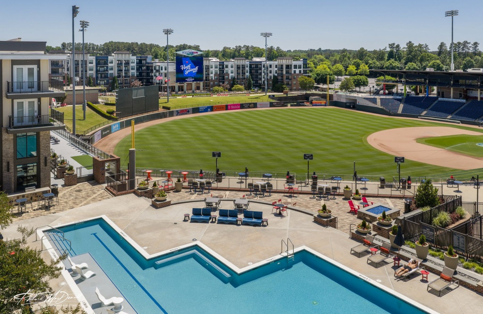 A baseball stadium sits behind a pool area, with Fieldhouse Apartments and trees in the background under a sunny sky.