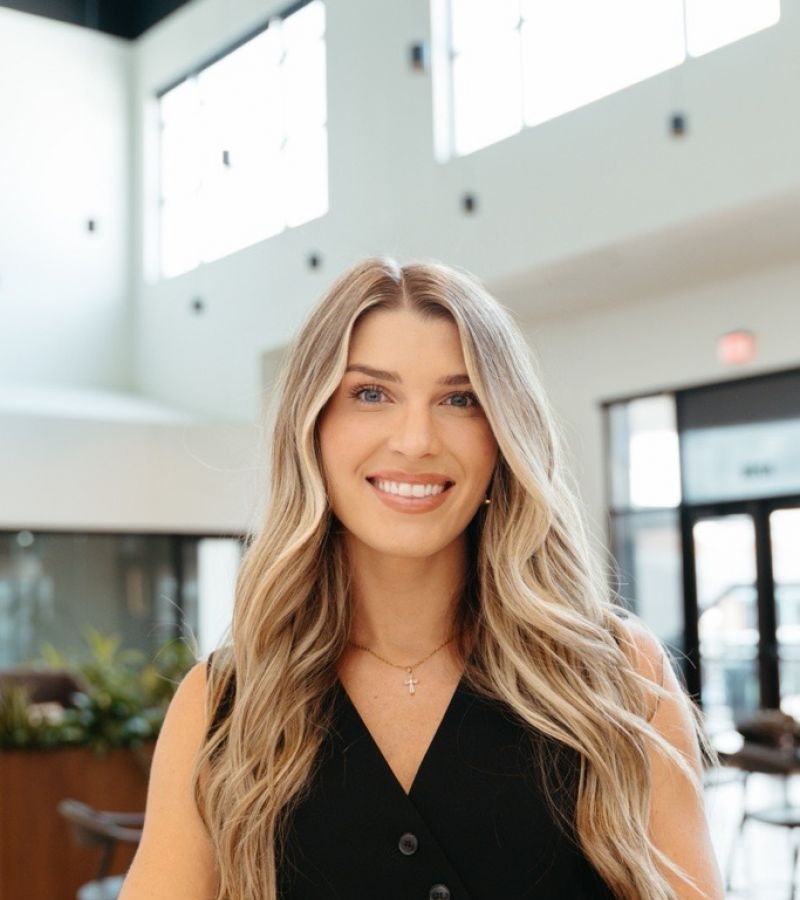 Woman with long blonde hair in a black sleeveless top smiling indoors by a table, photographed by Dev McDowell.