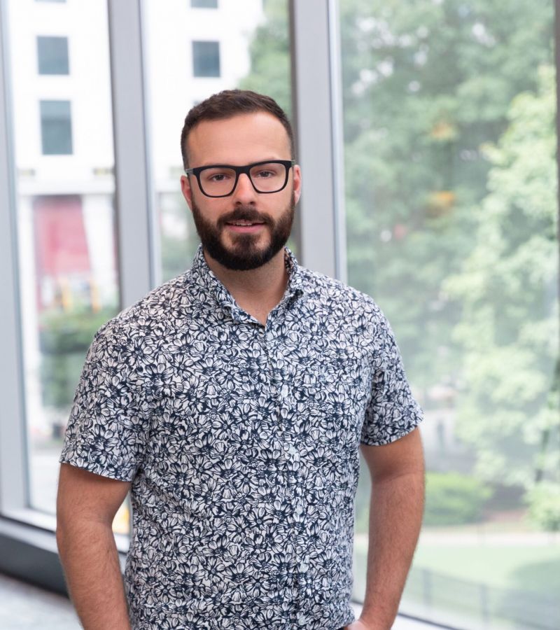 Man with glasses in a patterned shirt stands by large windows with a view of trees and buildings.
