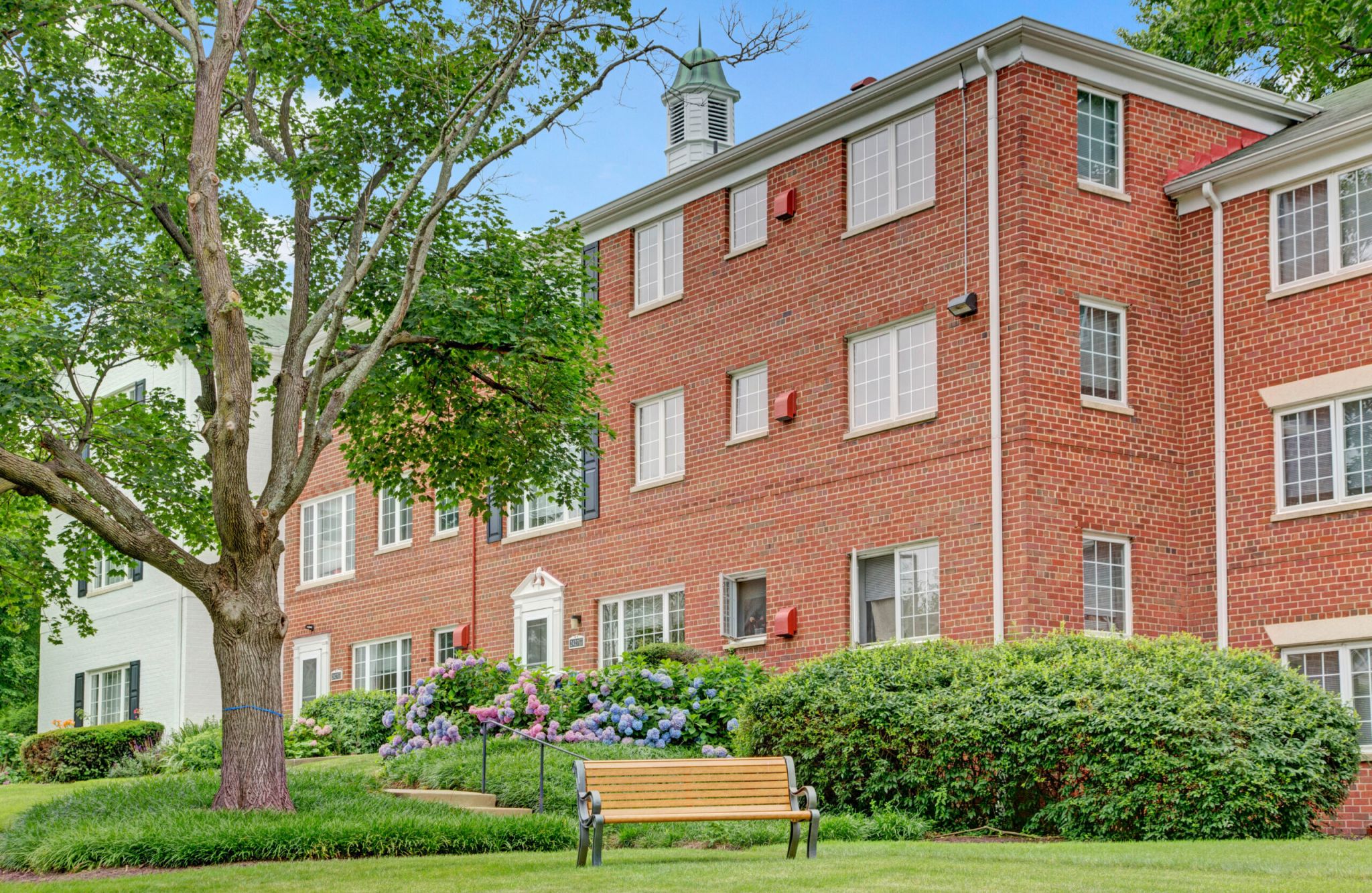 A red brick Braddock Lee apartment building with a garden and a bench in the foreground.