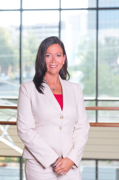 Woman in a cream suit standing indoors with large windows and trees in the background.