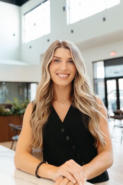 Woman with long blonde hair in a black sleeveless top smiling indoors by a table, photographed by Dev McDowell.