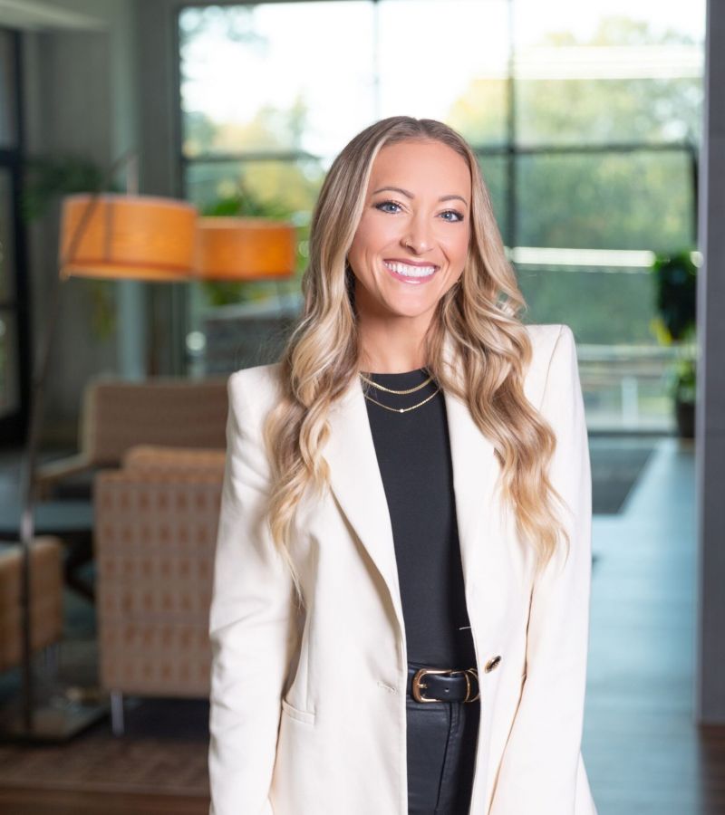 Smiling person in a white blazer and black outfit standing indoors with soft lighting and modern decor.