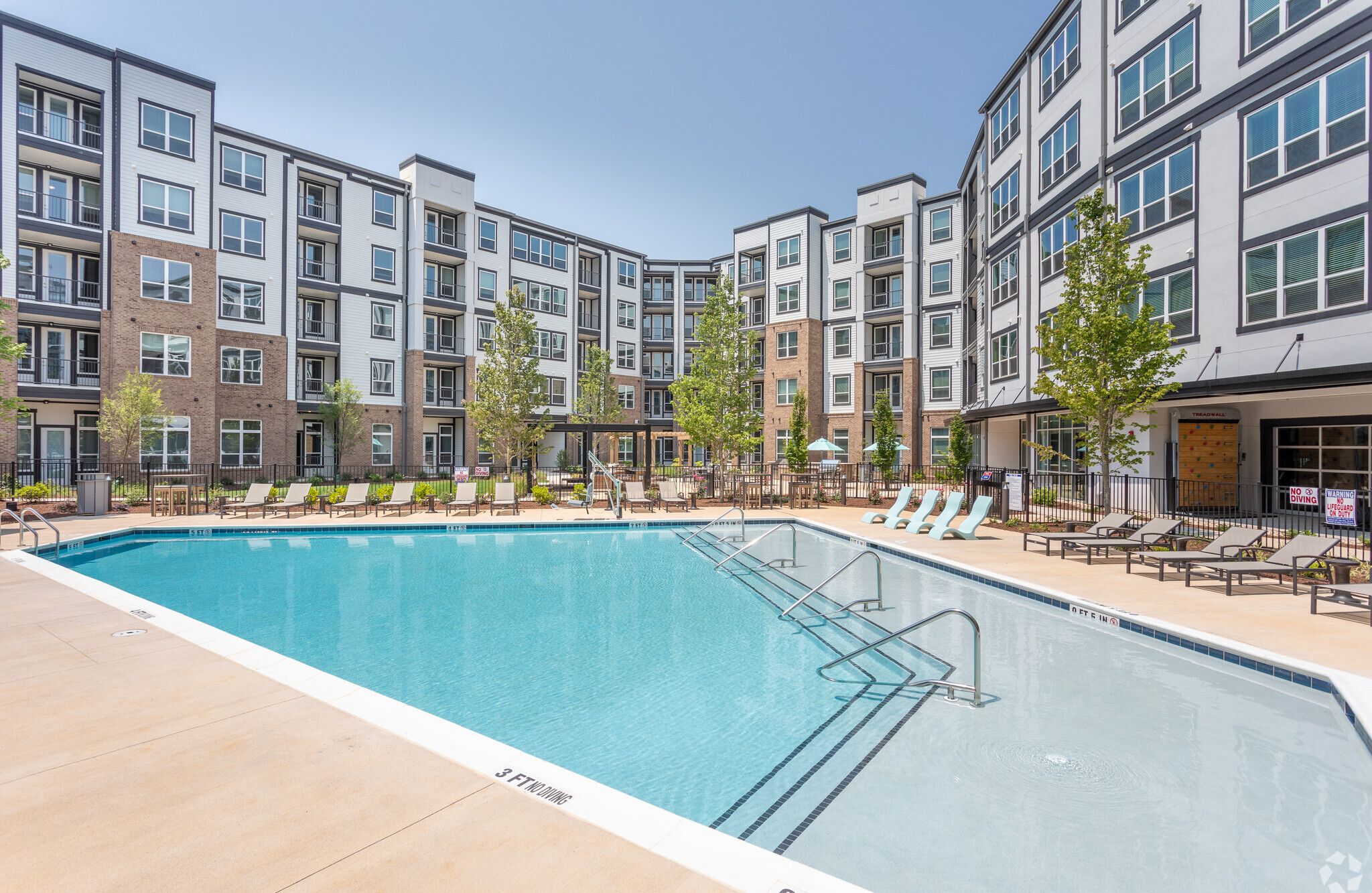 Large outdoor pool surrounded by lounge chairs and modern apartment buildings on a sunny day.